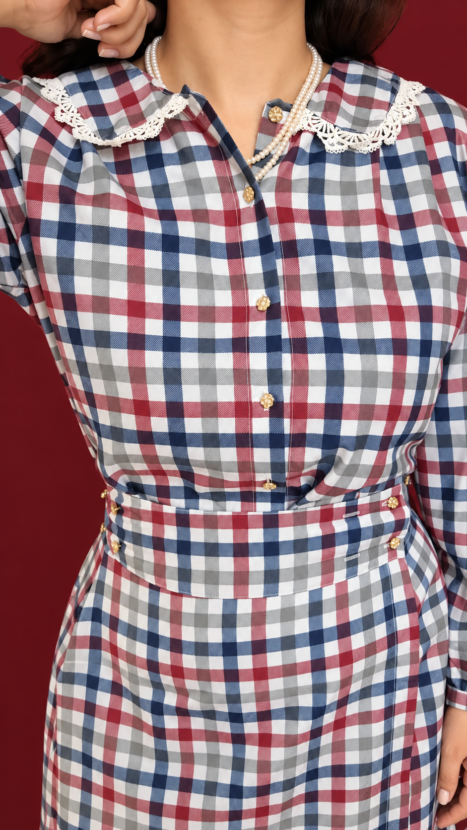A close-up of a person wearing a long-sleeve plaid dress with a red, navy, gray, and white check pattern, featuring a white lace Peter Pan collar and gold-tone buttons.