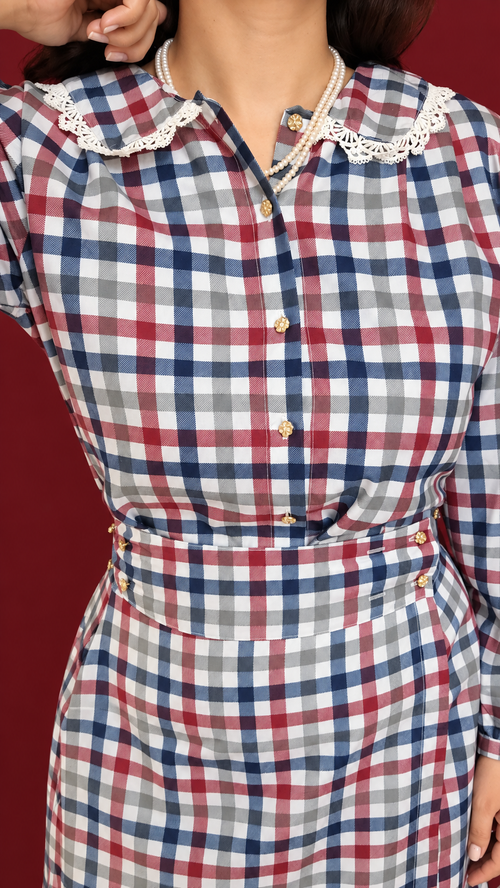 A close-up of a person wearing a long-sleeve plaid dress with a red, navy, gray, and white check pattern, featuring a white lace Peter Pan collar and gold-tone buttons.